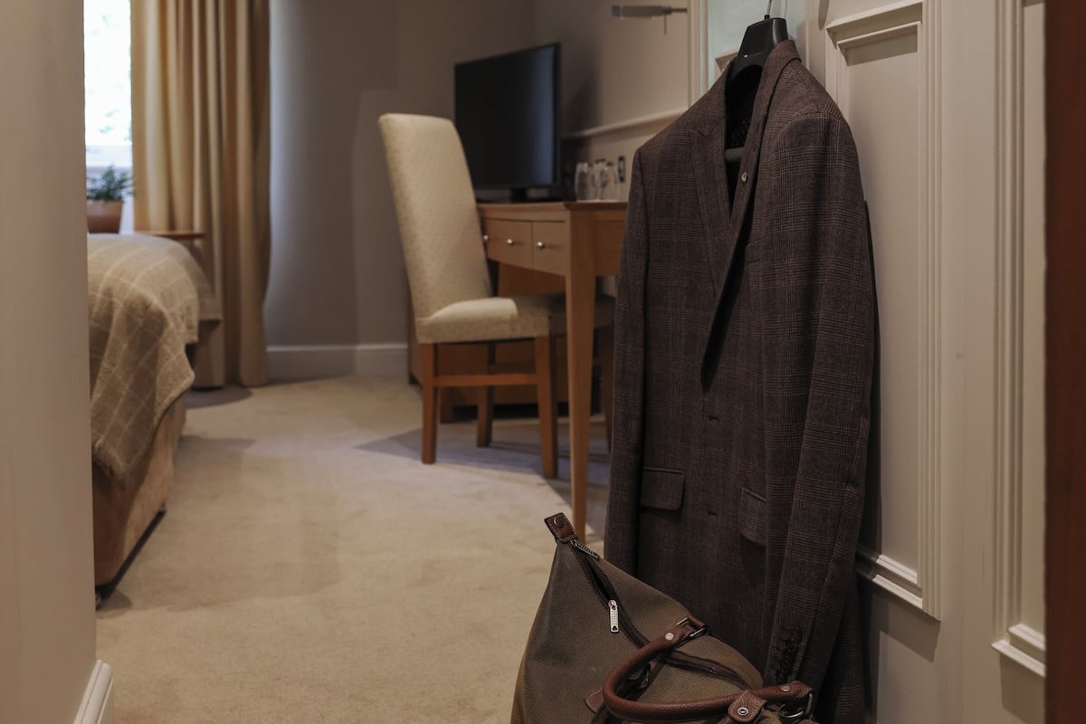 Habitación de hotel con cama, una silla, un escritorio con televisor y un perchero con abrigo y bolso.