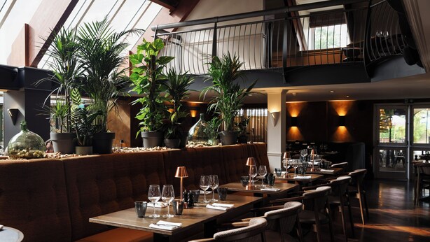 A restaurant with a high ceiling, wooden beams, and a mezzanine level. Tables are set with wine glasses and plates, and there are potted plants on the bar area. - Donnington Hotel, Spa & Golf (Newbury)