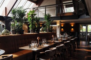 A restaurant with a high ceiling, wooden beams, and a mezzanine level. Tables are set with wine glasses and plates, and there are potted plants on the bar area. - Donnington Hotel, Spa & Golf (Newbury)