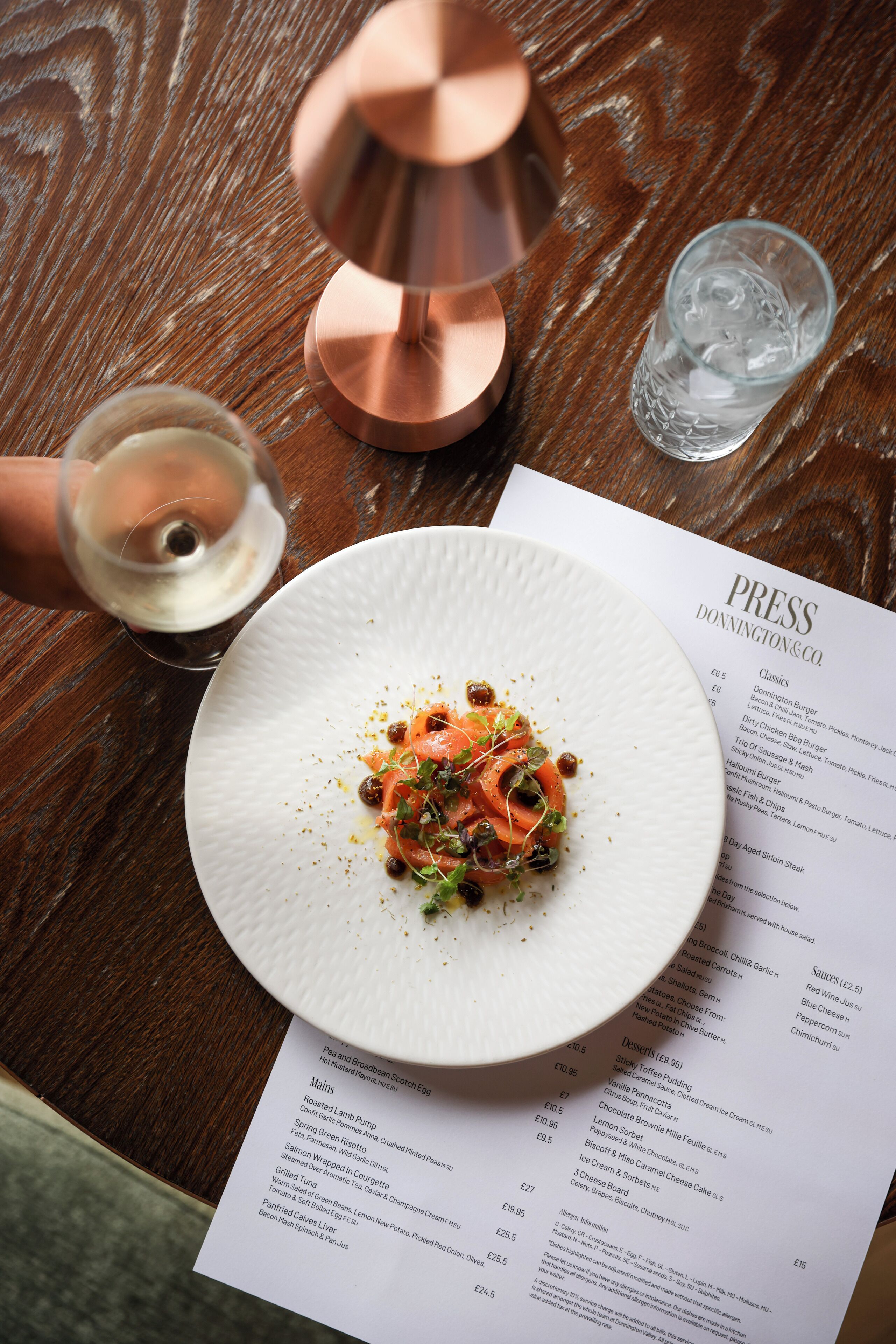 A menu with various dishes listed, a white plate with food, a glass of white wine, and a copper candle holder on a wooden table.