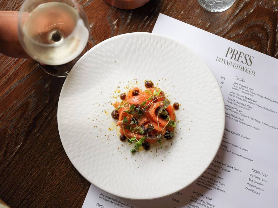 A menu with various dishes listed, a white plate with food, a glass of white wine, and a copper candle holder on a wooden table.