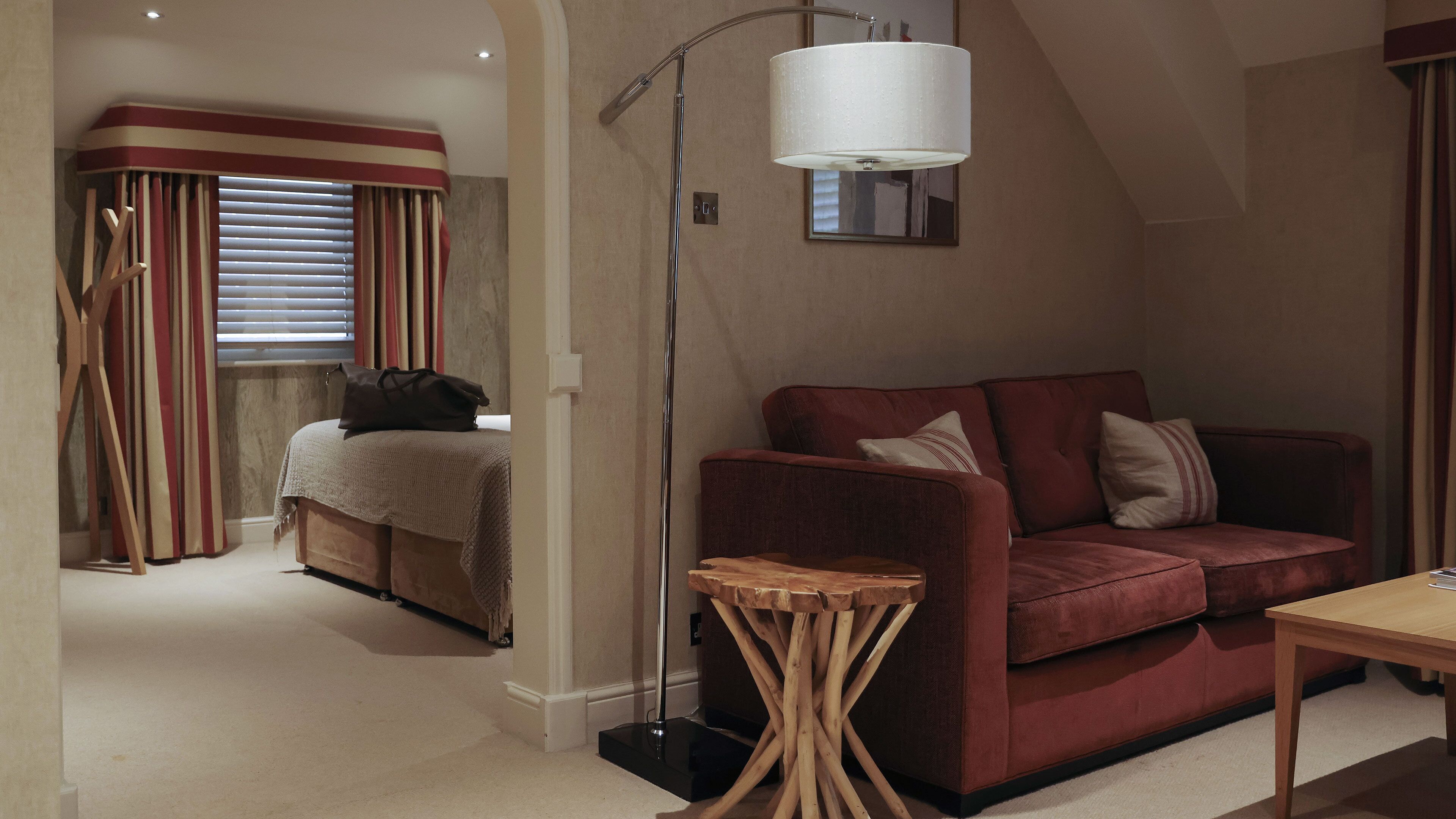 A modern hotel room with a red sofa, a small wooden side table, a floor lamp, and a bed with striped curtains.