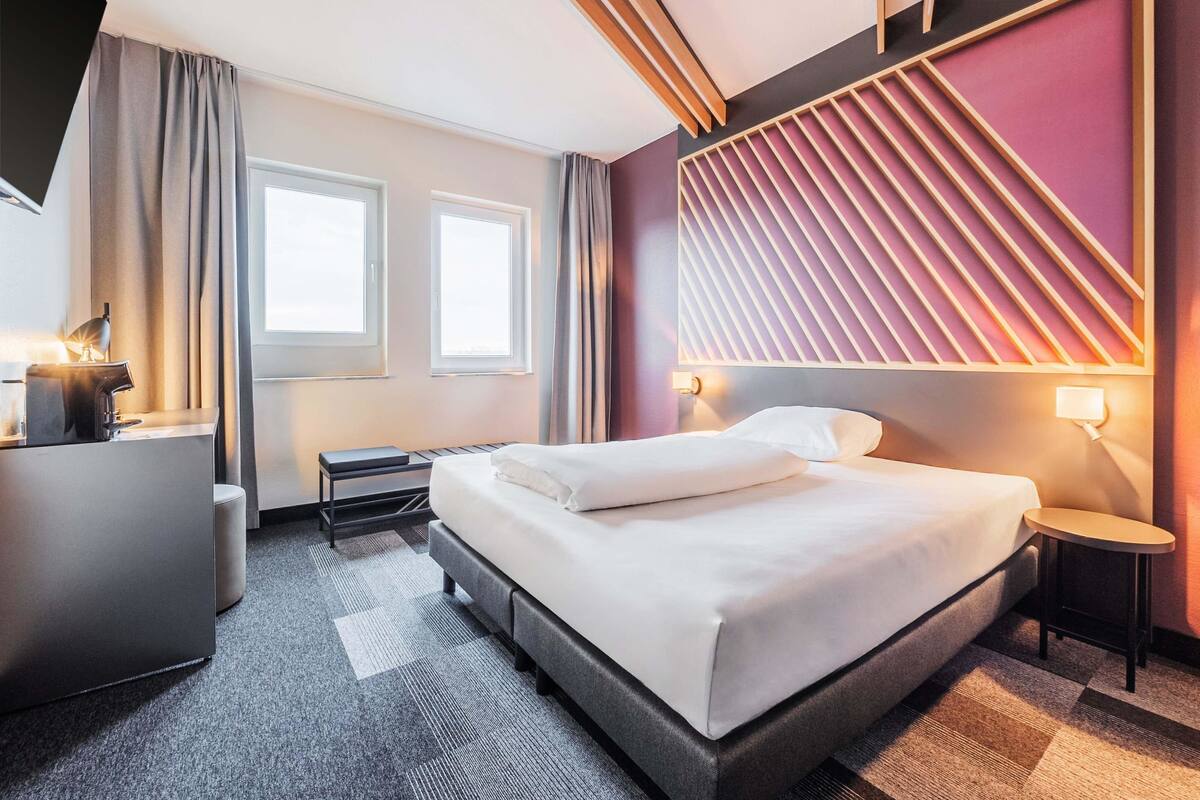 Une chambre d’hôtel moderne avec un grand lit, un bureau avec une lampe, une petite table et un luminaire fixé au mur.