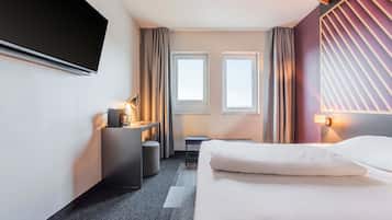 Habitación de hotel con cama, escritorio con lámpara, dos ventanas y televisor montado en la pared.