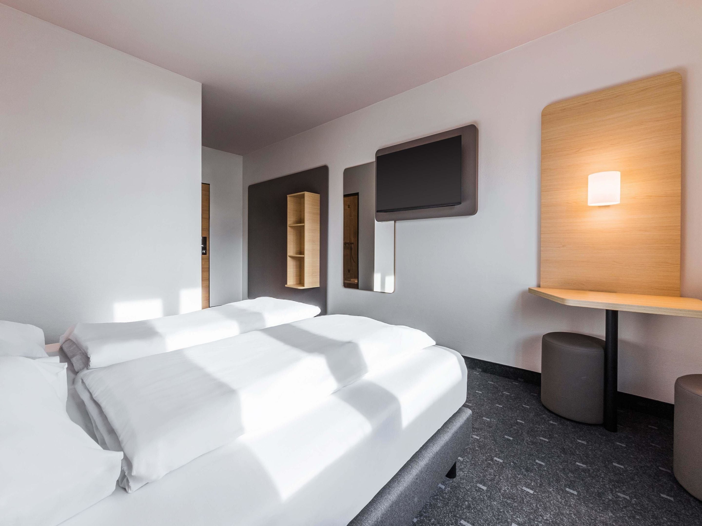 A modern hotel room with a large bed, a small desk, a TV mounted on the wall, and a shelf with a lamp.