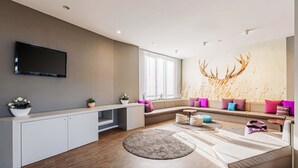 A modern living room with a large flat-screen TV, a sofa with colorful cushions, a round coffee table, and a decorative wall piece. - B&B HOTEL Rastatt (Rastatt)
