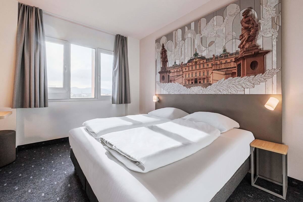 Une chambre d’hôtel avec un grand lit, une fenêtre avec des rideaux, une œuvre d’art accrochée au mur et une table de chevet.