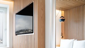 A modern hotel room with a wooden slat wall design, a large mirror, and a bed with a view of a cityscape through a window.