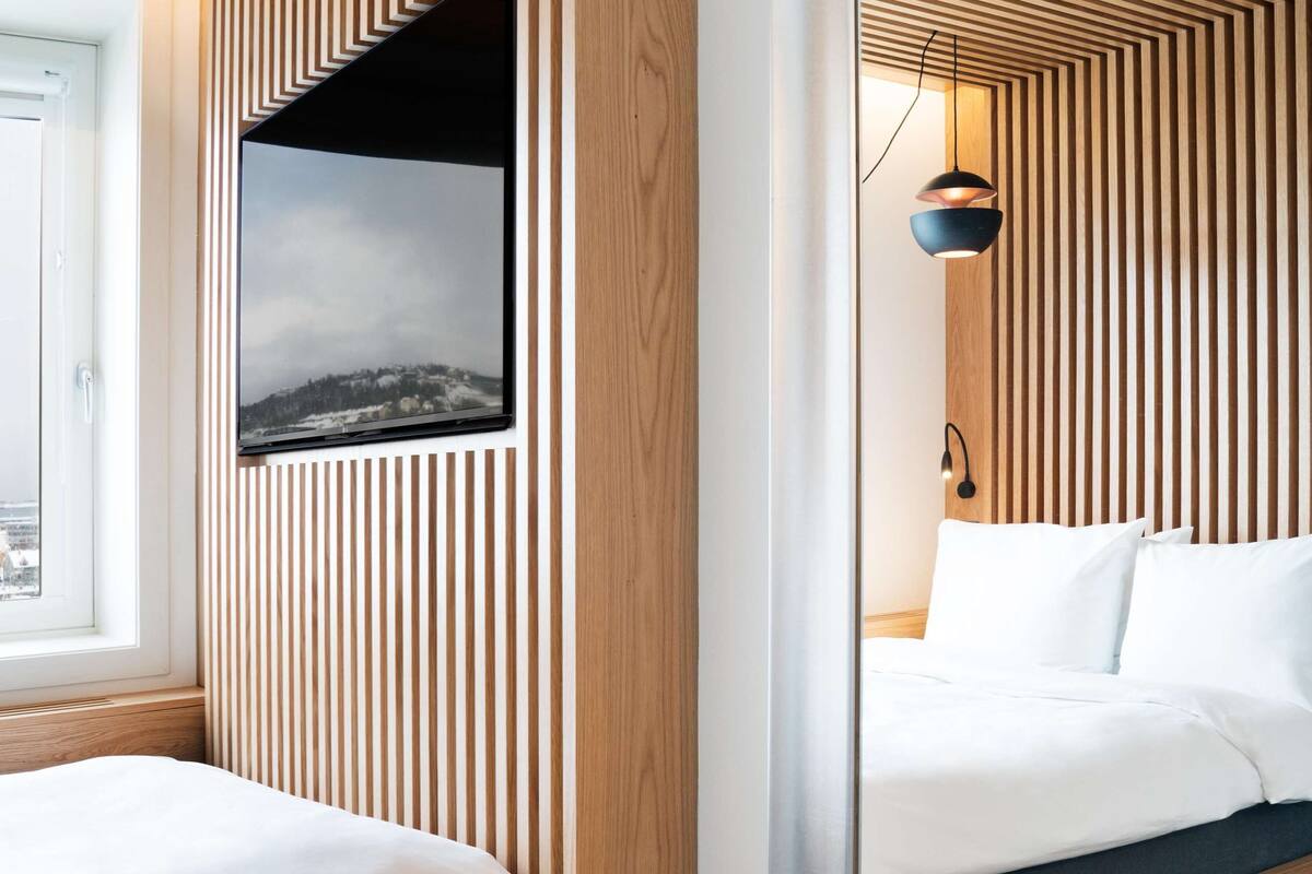 A modern hotel room with a wooden slat wall design, a large mirror, and a bed with a view of a cityscape through a window.