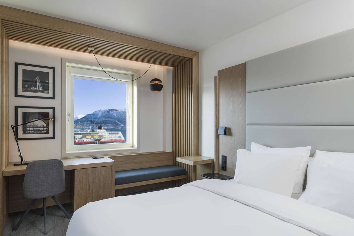 A modern hotel room with a large bed, a desk with a chair, a window with a view of mountains and a ship, and a wooden accent wall.