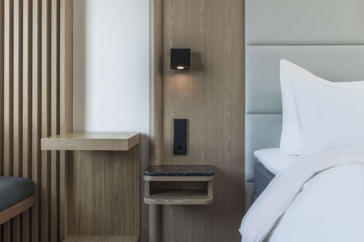 A modern hotel room with a wooden accent wall, a built-in shelf, a bedside table, and a bed with white bedding.