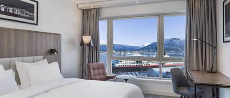 A hotel room with a large bed, a desk, a chair, and a view of a city and mountains through a large window.