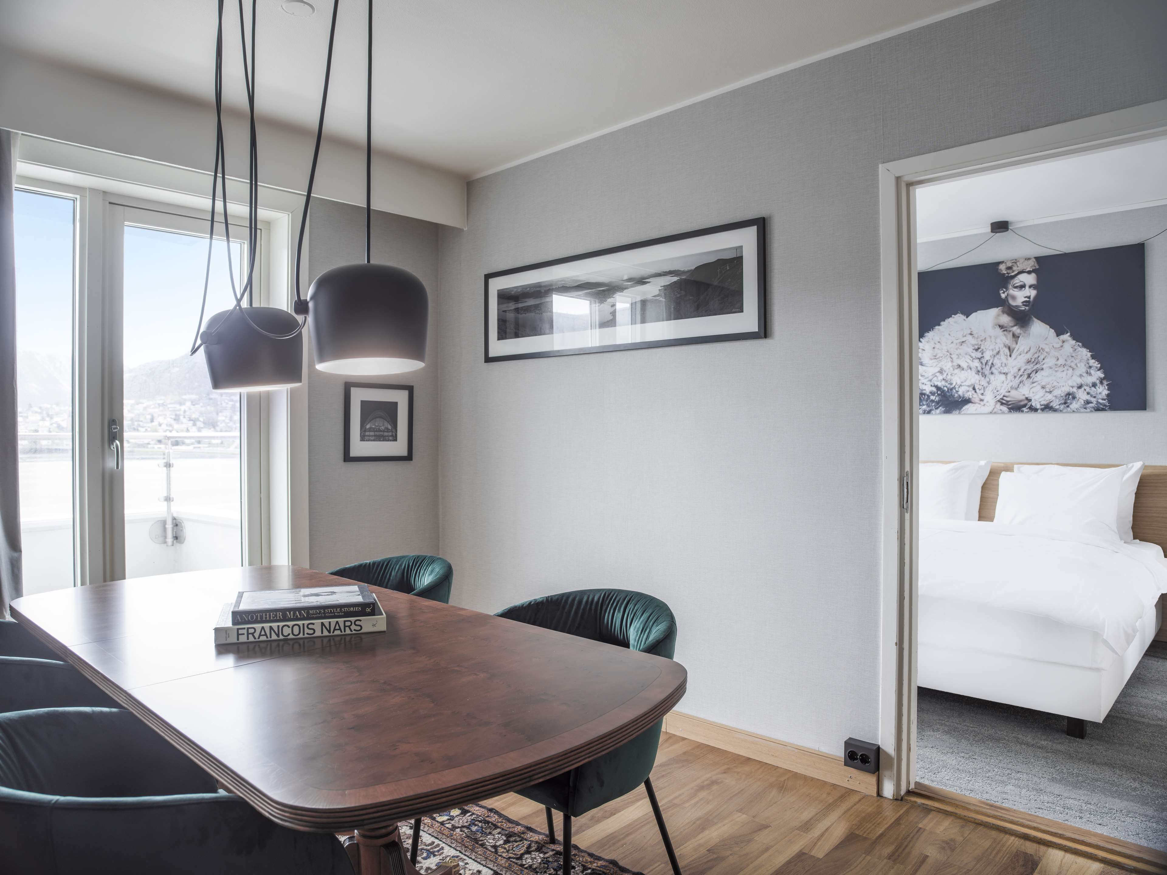 Una habitación de hotel moderna con una mesa de comedor de madera, una silla verde, una cama con ropa blanca y un amplio ventanal con vista a la ciudad.