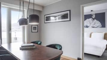 Una habitación de hotel moderna con una mesa de comedor de madera, una silla verde, una cama con ropa blanca y un amplio ventanal con vista a la ciudad.