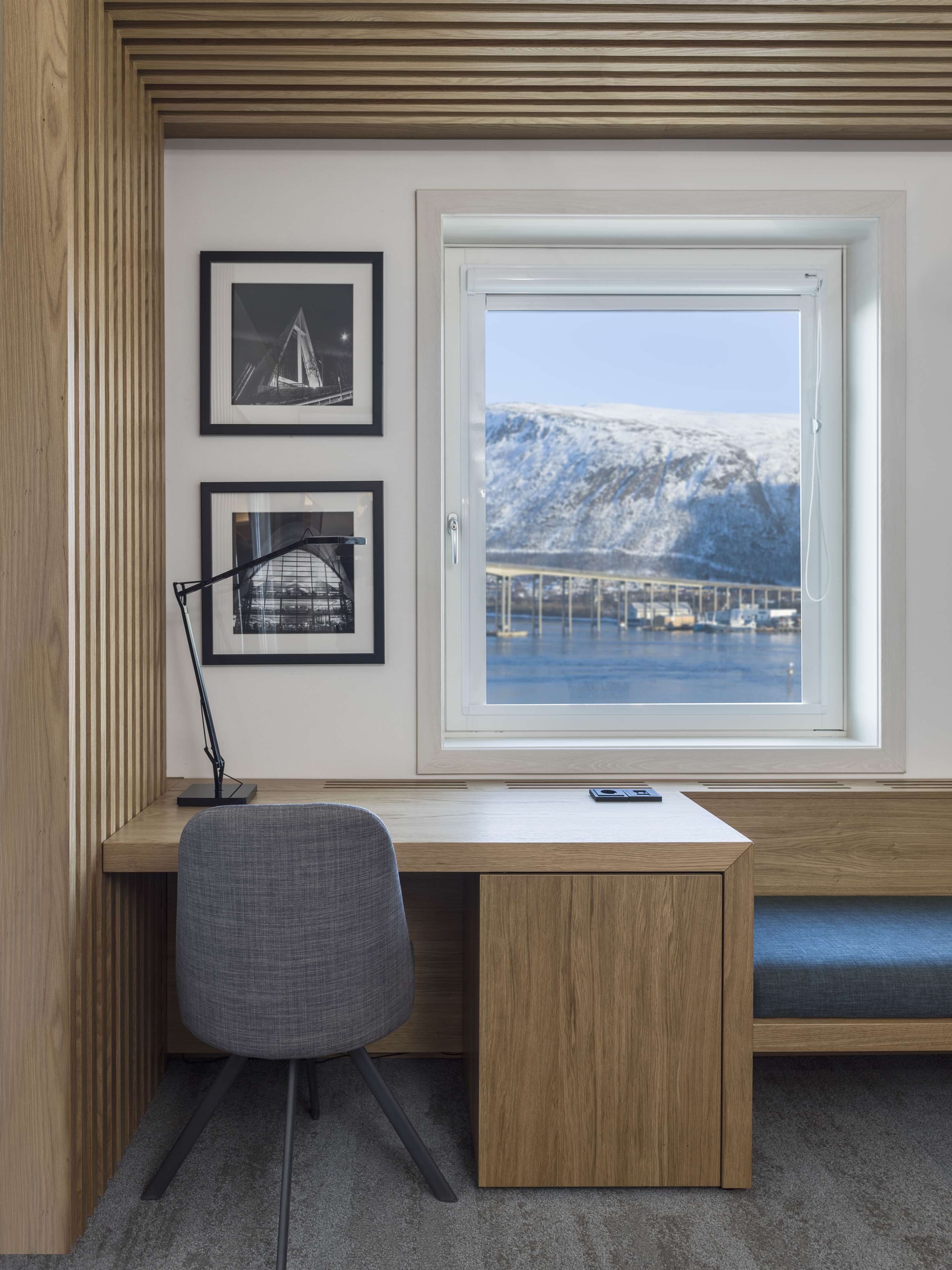 Un espacio de trabajo moderno con escritorio, silla y un amplio ventanal que ofrece vistas a un paisaje nevado y un puente.