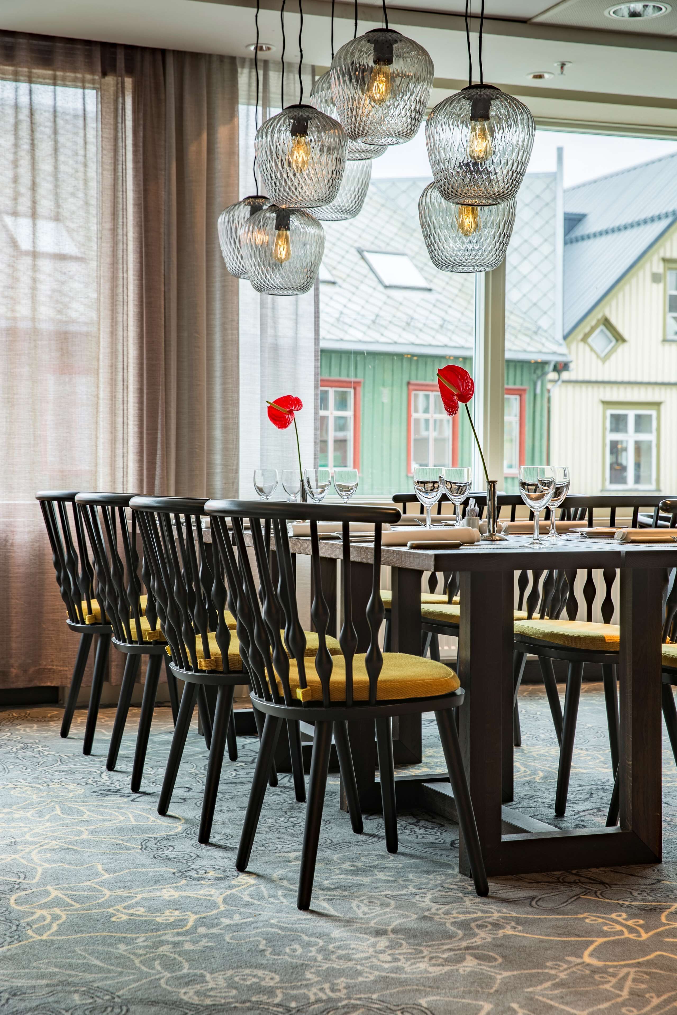 A dining area with a table set for four, featuring black chairs with yellow cushions, a patterned carpet, and a view of buildings outside.