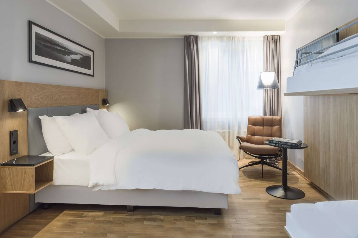 A hotel room with a bed, bedside table, lamp, chair, and a small table with books.