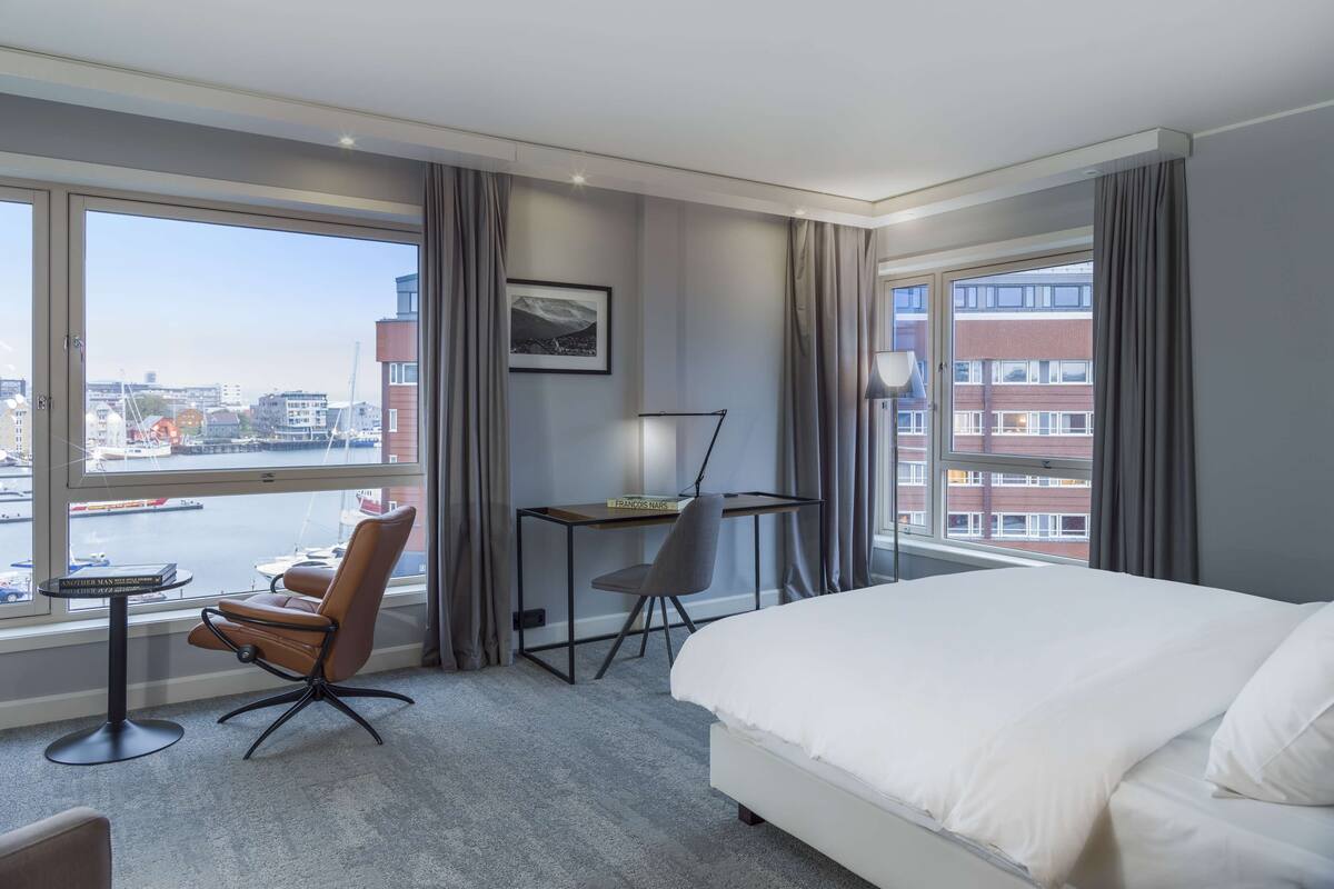 A hotel room with a large bed, a desk, a chair, and a view of the city through large windows.