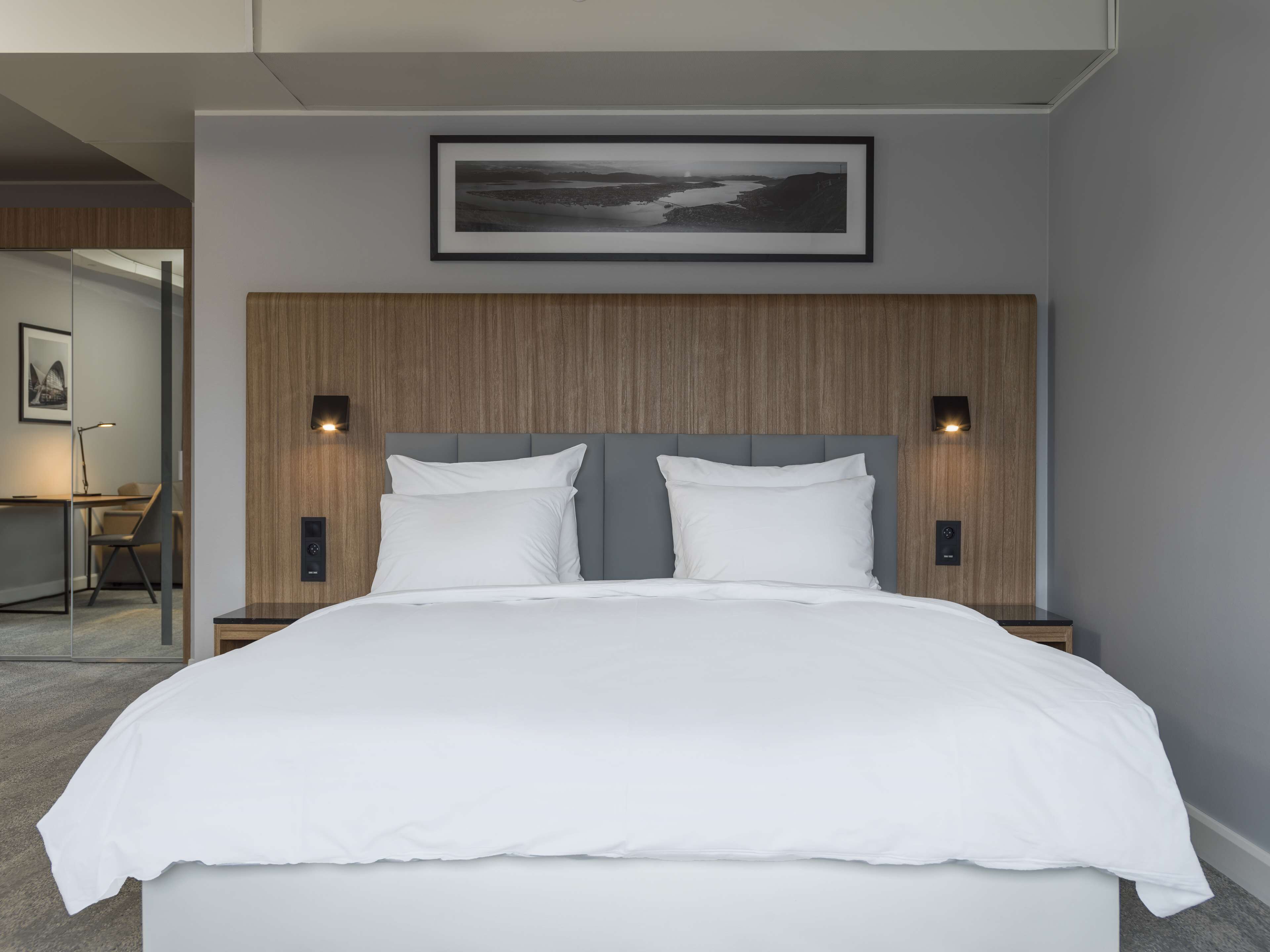 A modern bedroom with a large bed, wooden headboard, two bedside lamps, and a framed landscape picture above the bed.