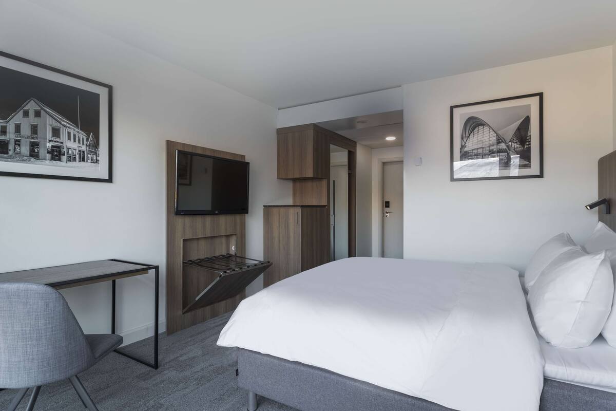 A modern hotel room with a bed, a desk, a TV, and two framed pictures on the walls.