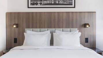 Habitación de hotel moderna con cabecera de madera, dos almohadas blancas y una fotografía enmarcada de un paisaje urbano sobre la cama.