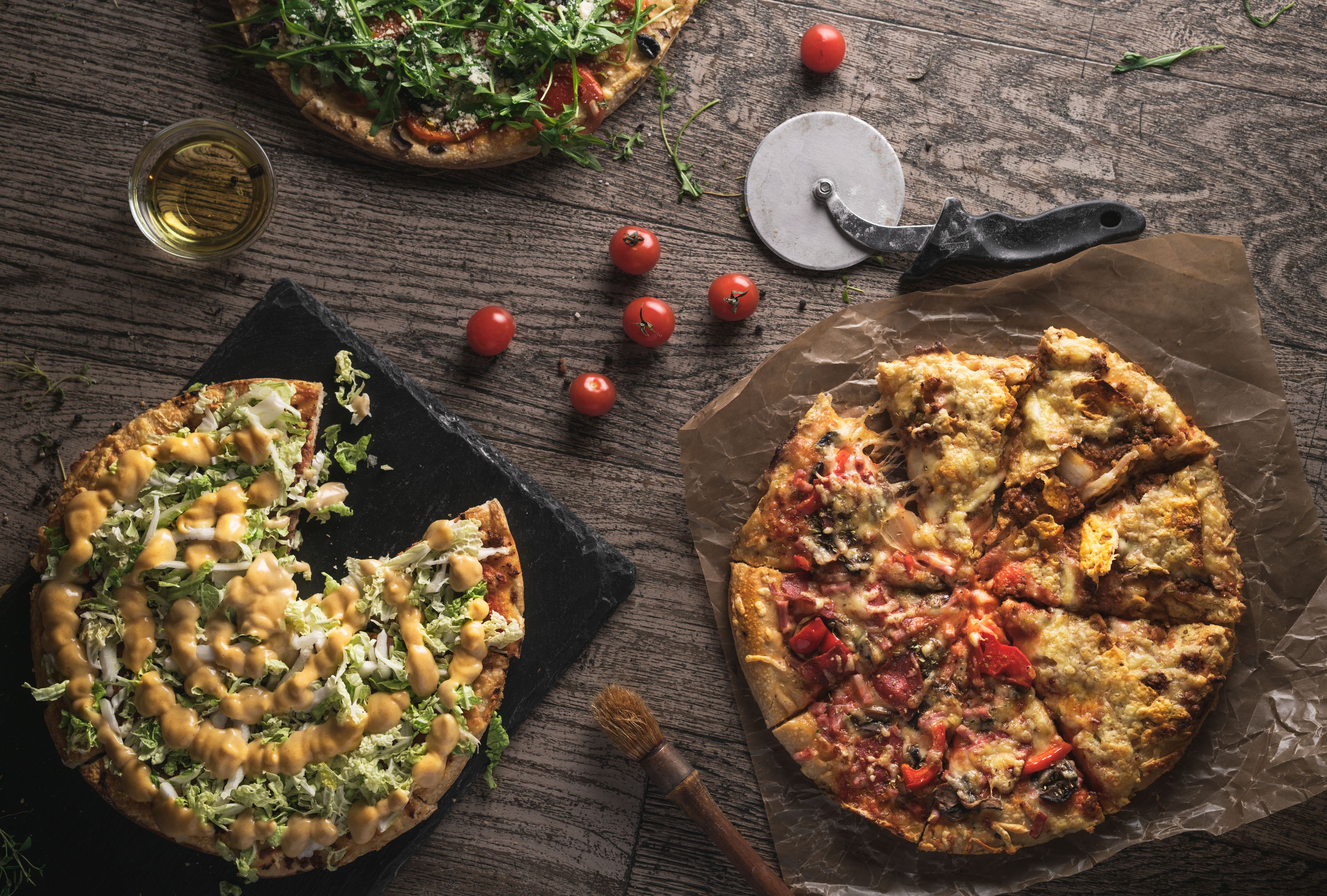Two pizzas on wooden surfaces, one cut into slices and the other whole, with a pizza cutter and tomatoes nearby.