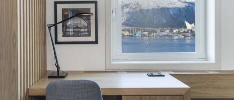 A modern room with a wooden desk, a grey chair, and a large window with a view of a snowy mountain and a bridge.