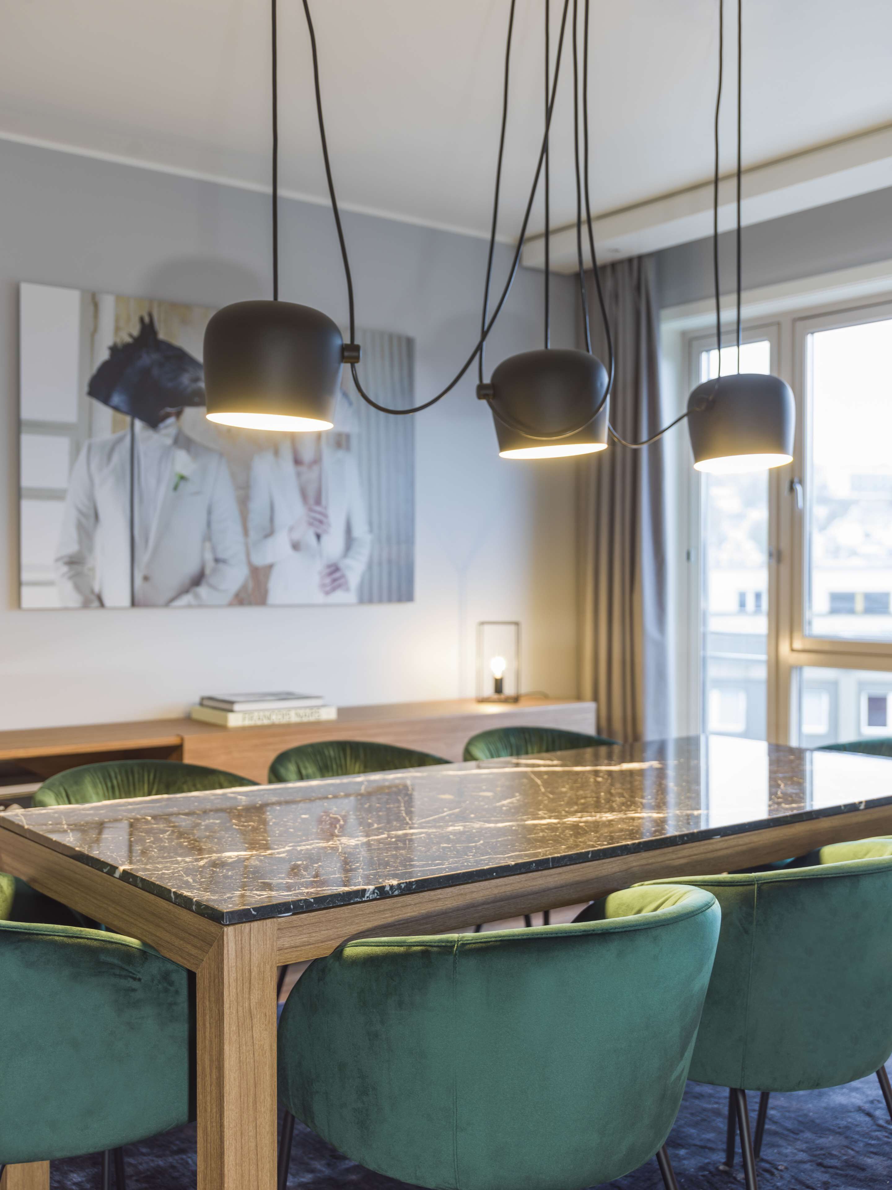 Zona pranzo moderna con tavolo in vetro, sedie imbottite verdi e illuminazione a sospensione.