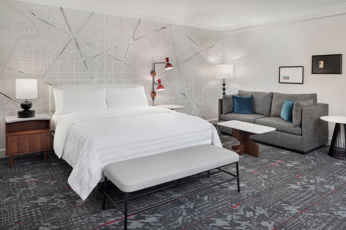 A modern hotel room with a large bed, a grey sofa, a small table, and a bench.