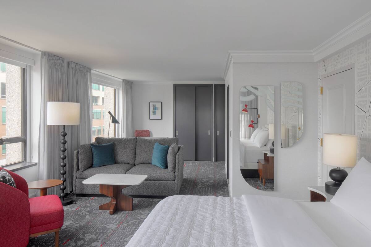 A hotel room with a bed, a sofa, a coffee table, a red armchair, a floor lamp, and a mirror.