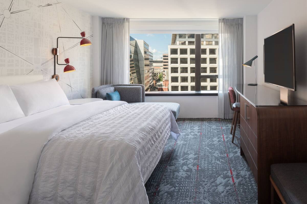 A hotel room with a large bed, a desk, a chair, a TV, and a view of the city through the window.