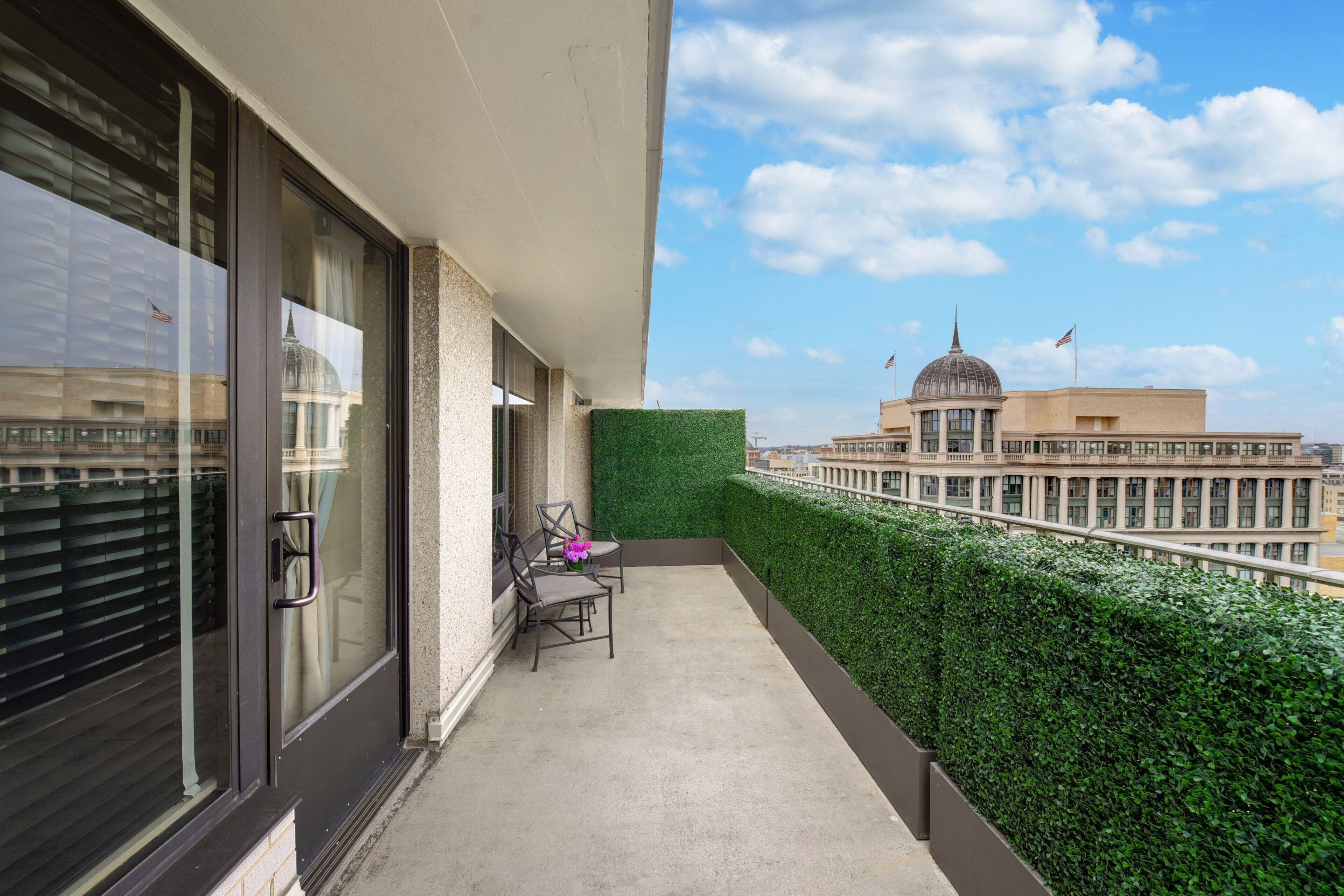 A balcony with a view of a building with a dome and flags, a green hedge, and a chair with a vase of flowers.