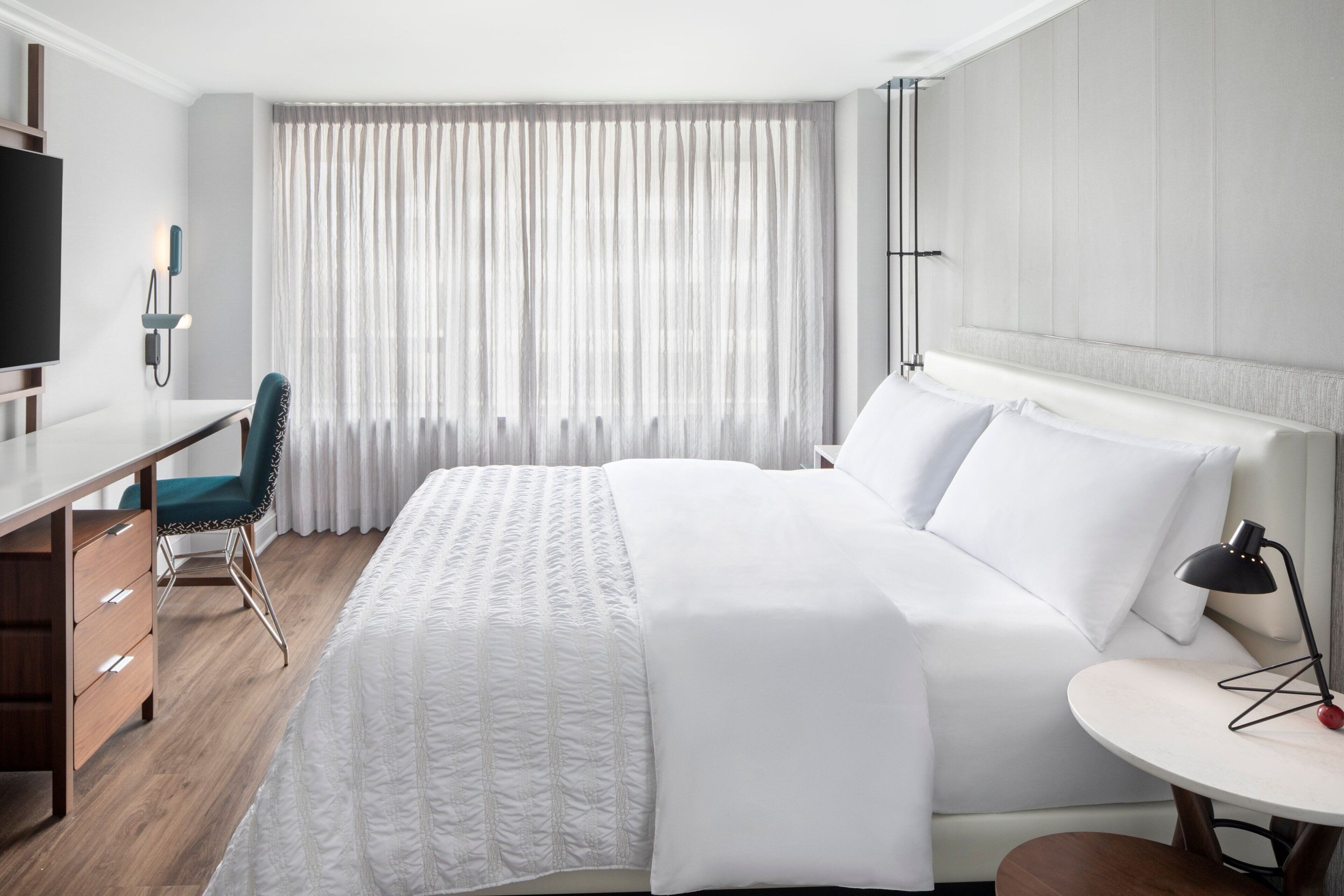 A hotel room with a bed, a desk, a chair, a TV, and a window with curtains.