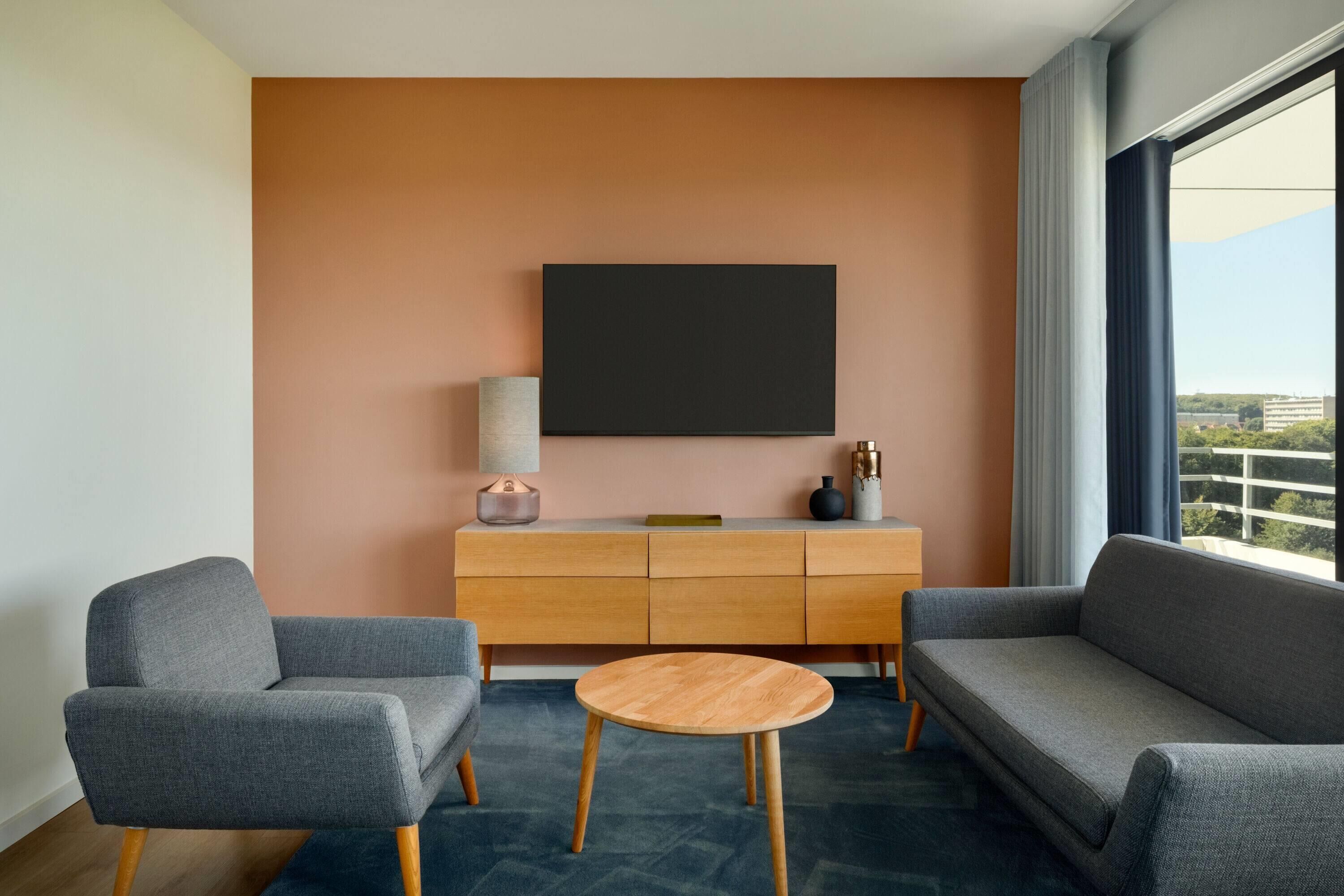 A modern hotel room with a grey sofa, a wooden coffee table, and a large TV on the wall.