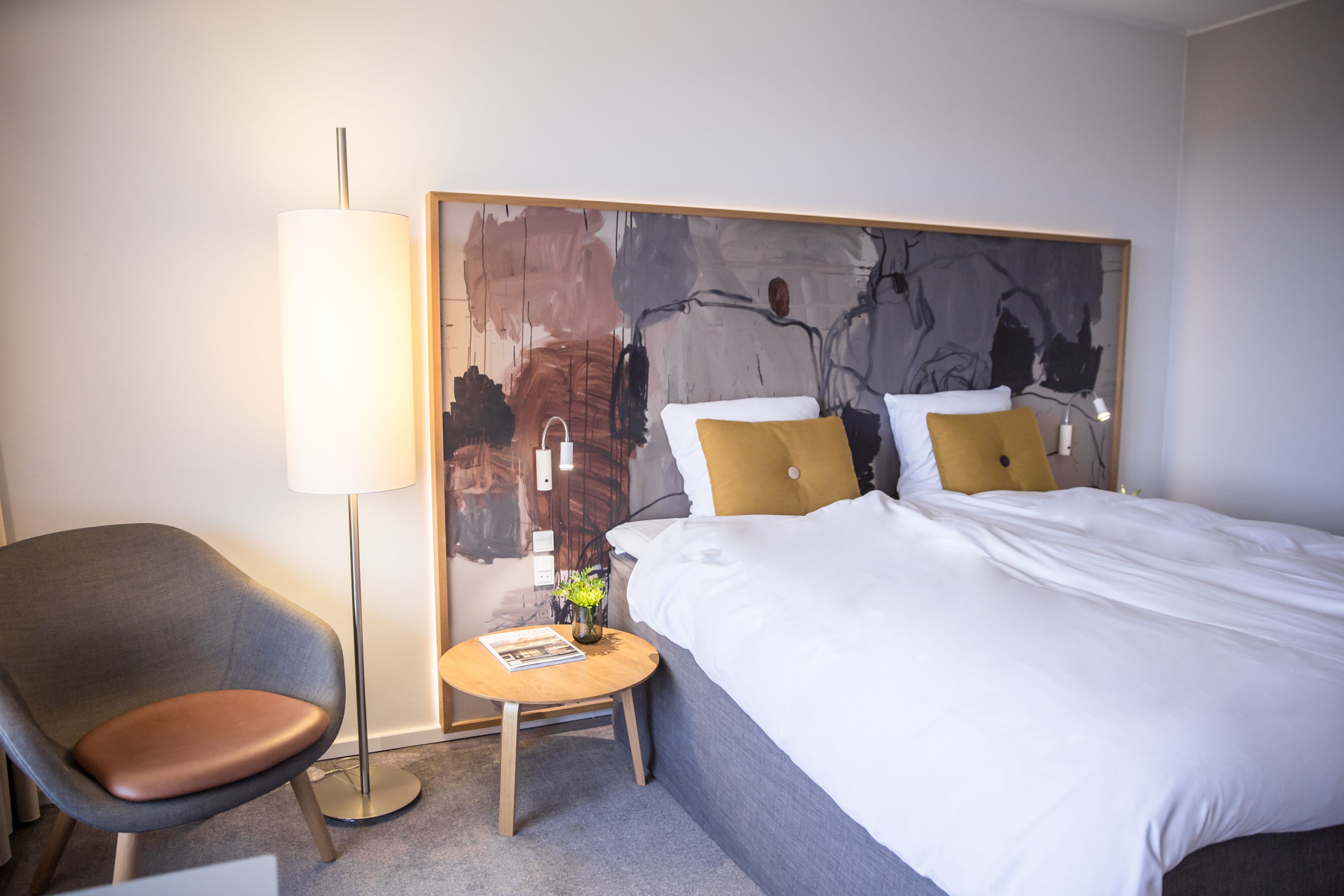A hotel room with a bed, a chair, a small table, a lamp, and a wall art piece.