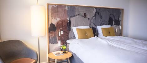 A hotel room with a bed, a chair, a small table, a lamp, and a wall art piece.
