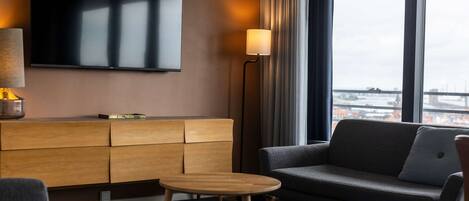 A modern hotel room with a large flat-screen TV mounted on the wall, a wooden console table with a lamp, a grey sofa, and a small round table.