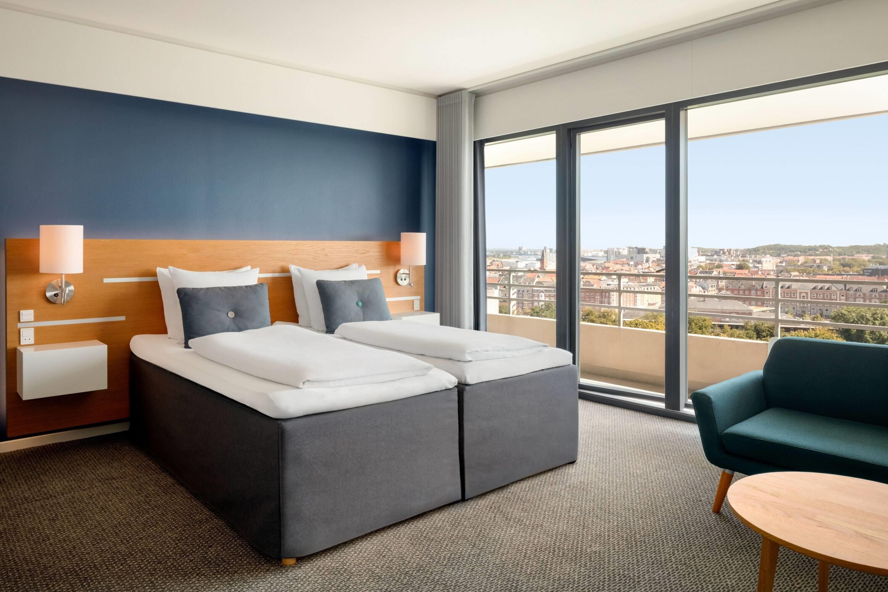 A hotel room with a bed, a sofa, a small table, and a large window offering a city view.
