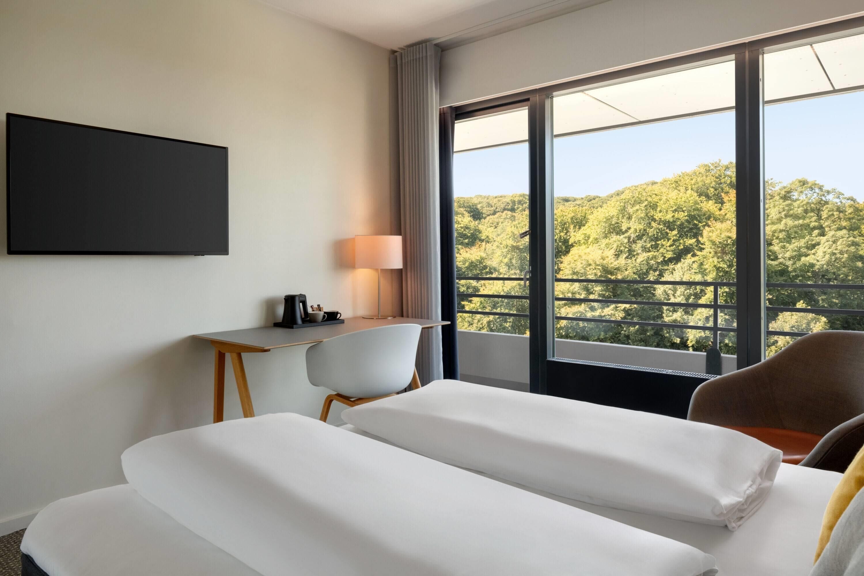 A hotel room with two beds, a desk, a chair, and a large window with a view of trees.