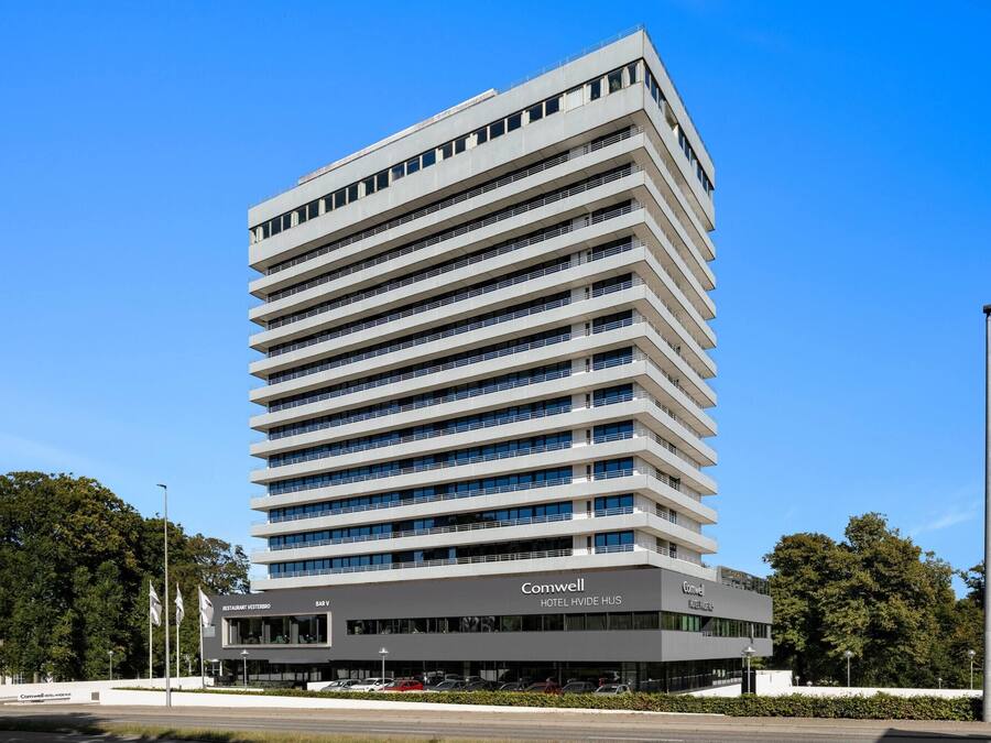 Un edificio moderno de hotel con varios pisos, techo plano y un cartel llamativo que dice 'Comwell Hotel Hvide Hus'.