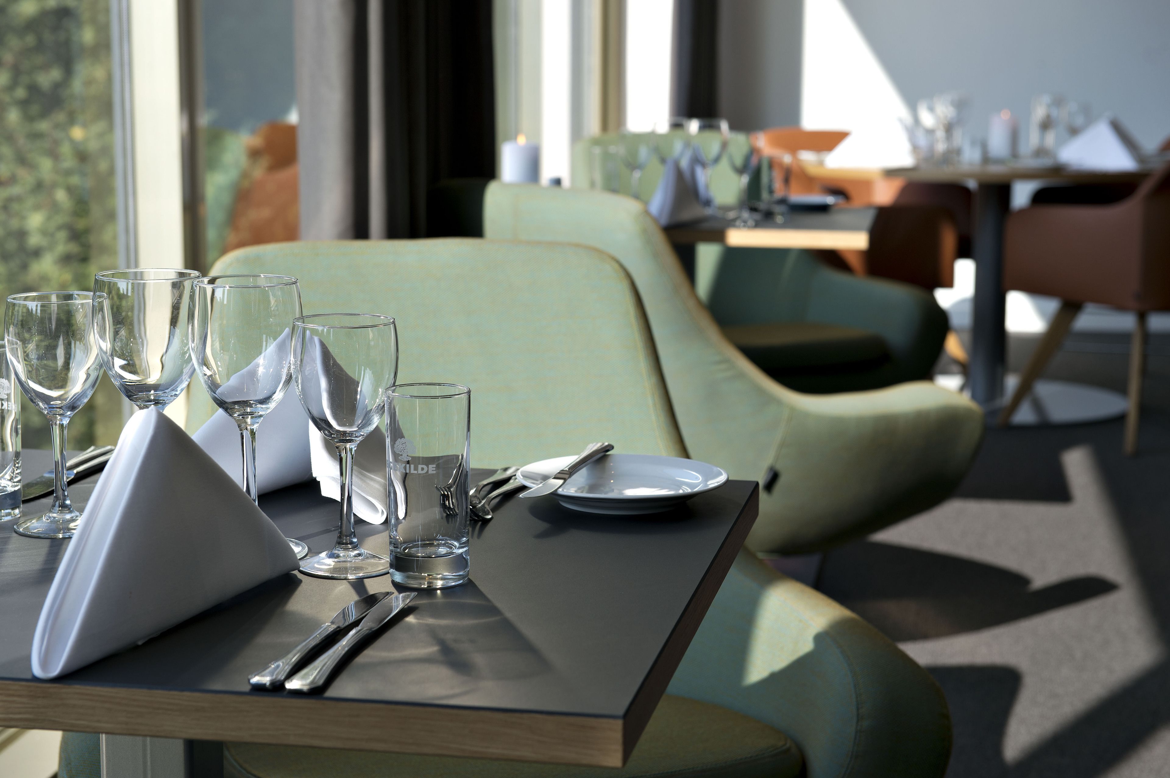 A dining table set with wine glasses, a plate, and cutlery in a modern restaurant with green upholstered chairs and a view of the outdoors.