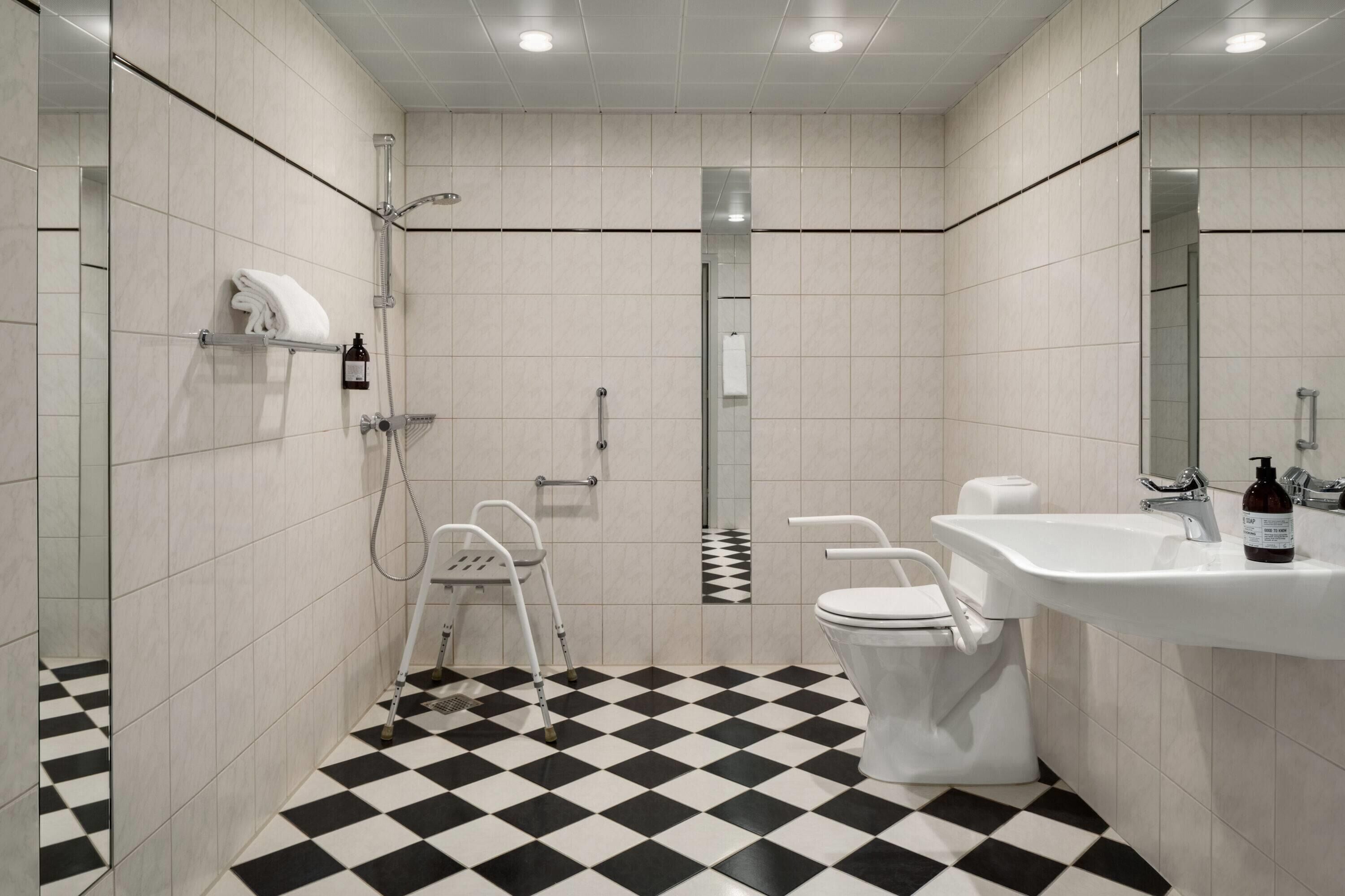Et moderne badeværelse med skakternet gulv, bruser, toilet, vask og spejl.