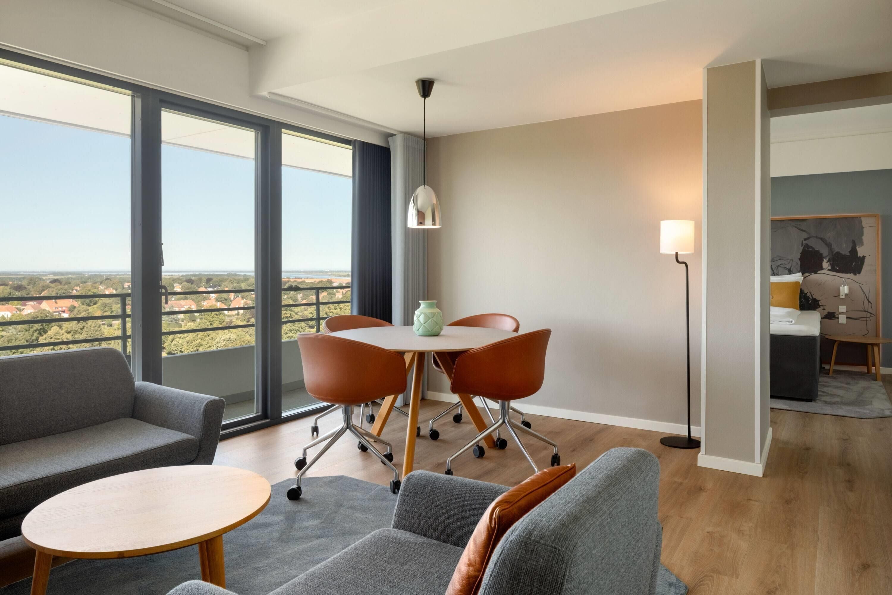 Ein modernes Wohnzimmer mit einer Couch, Sesseln, einem runden Tisch und einem Esstisch mit Stühlen. Ein großes Fenster bietet Blick auf die Stadtlandschaft.