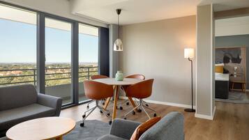 Ein modernes Wohnzimmer mit einer Couch, Sesseln, einem runden Tisch und einem Esstisch mit Stühlen. Ein großes Fenster bietet Blick auf die Stadtlandschaft.