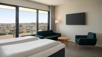 Habitación de hotel con un ventanal grande, dos sillones verdes, una mesita redonda y una cama con ropa blanca.