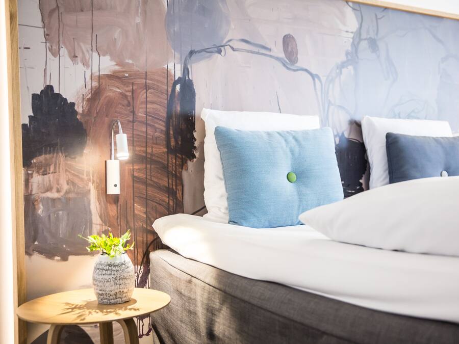 A modern hotel room with a bed, a bedside table with a vase, and a wall with abstract art.