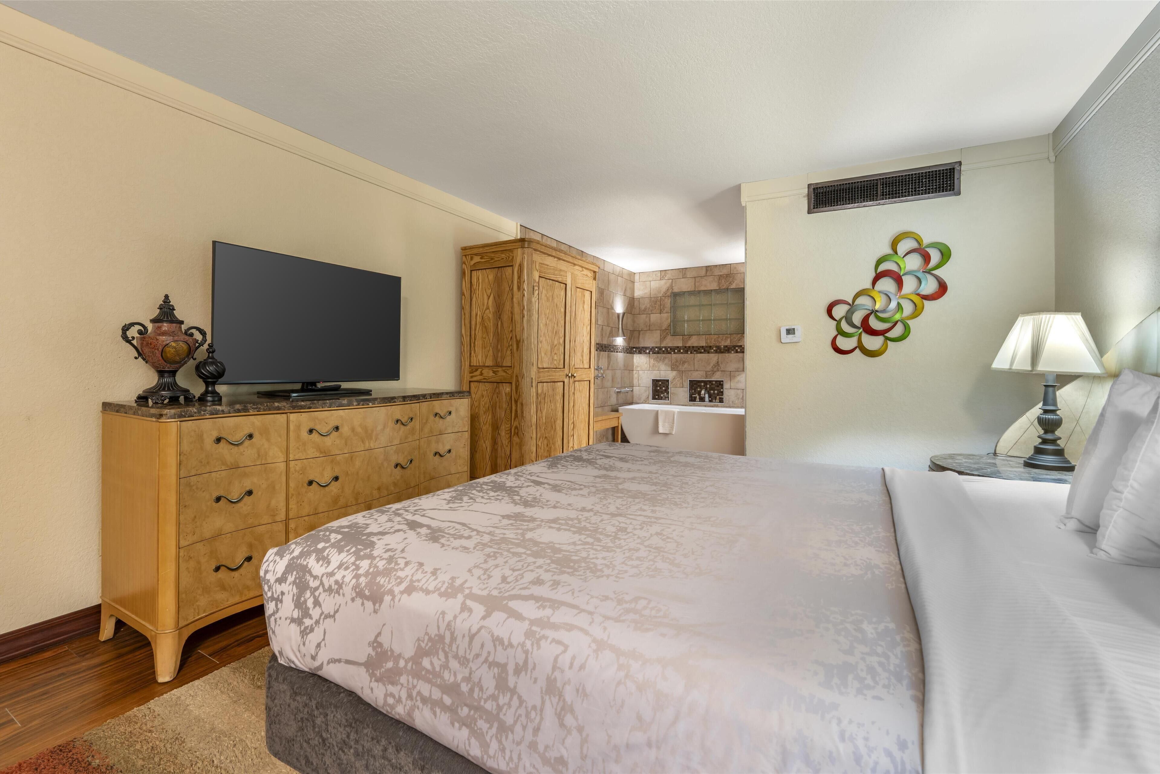 Un dormitorio con una cama, un aparador de madera, una televisión y un baño al fondo.