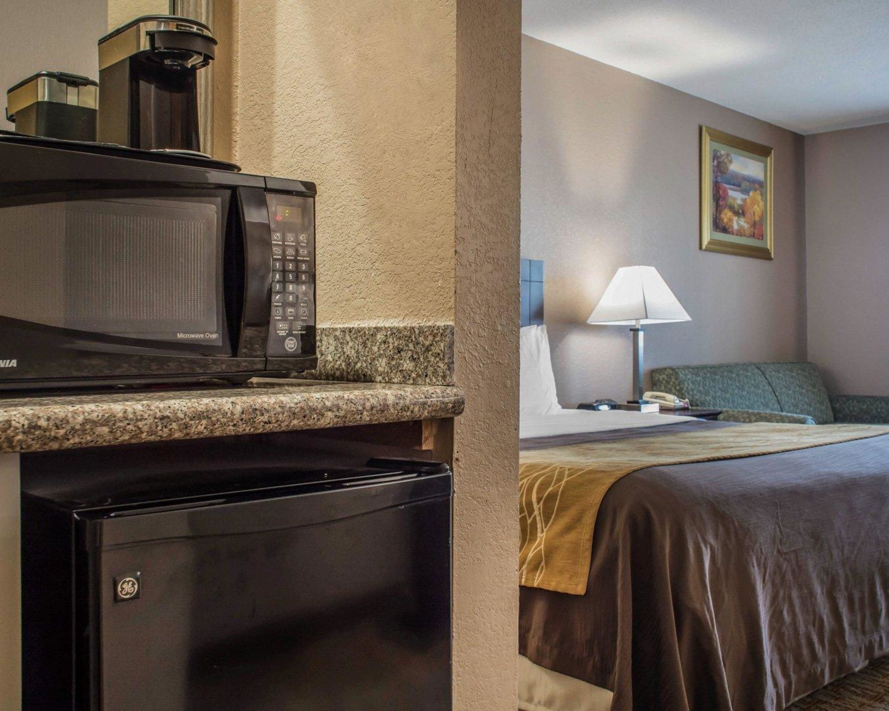 A hotel room with a microwave oven, a mini refrigerator, a bed with a green headboard, a bedside table with a lamp, and a painting on the wall.