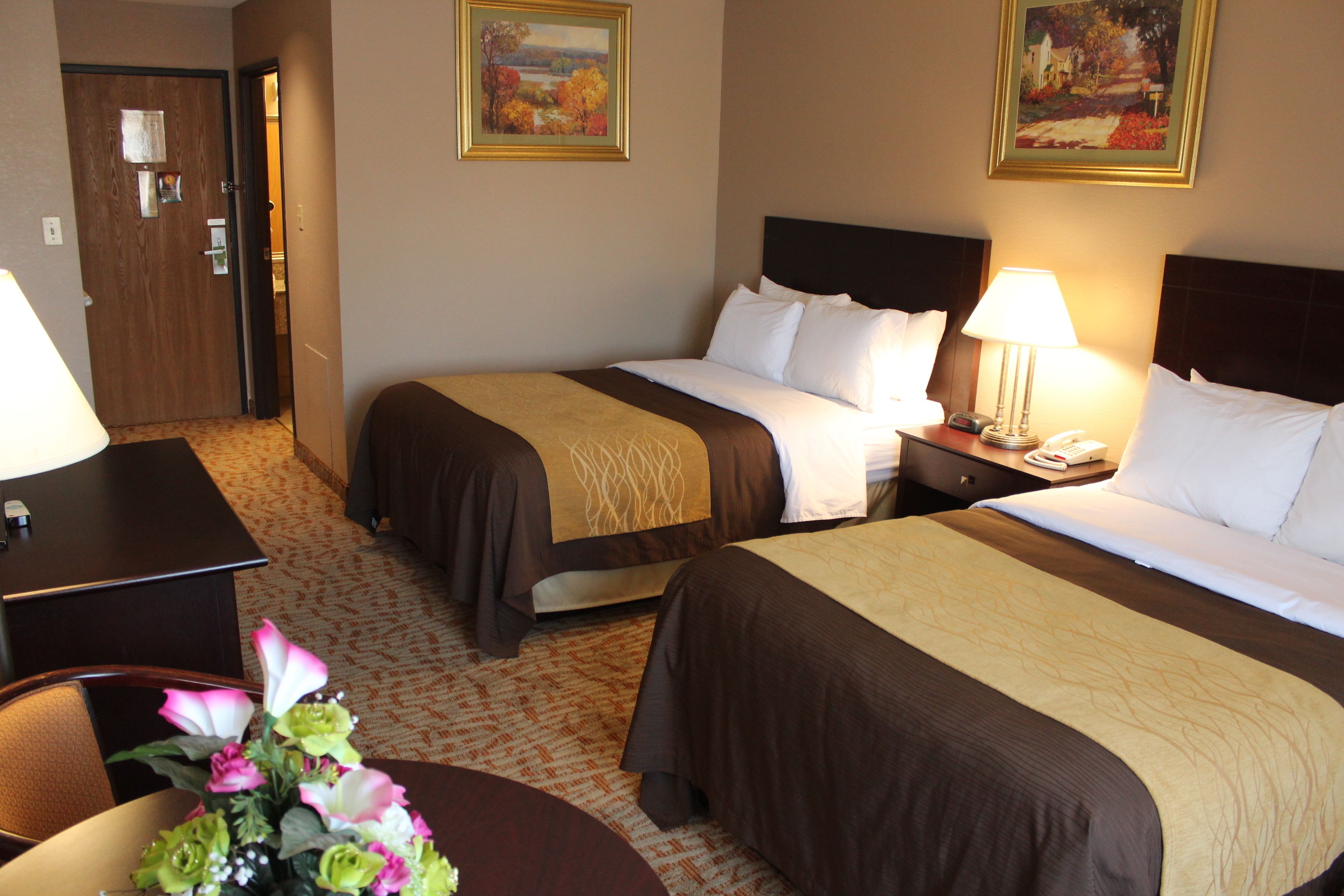 A hotel room with two beds, a desk, and a chair. There are paintings on the wall and a vase with flowers on the desk.