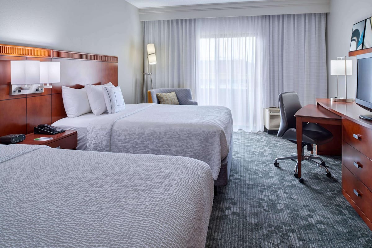 A hotel room with two beds, a desk, a chair, a TV, and a lamp.